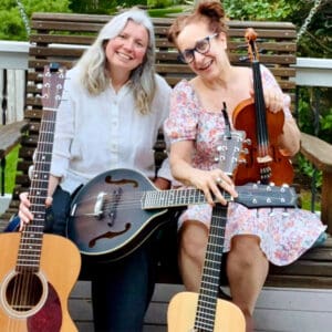 The Jamesons Duo sitting on a backporch swing holding guitars, fiddle and mandolin; Amy Glenney, left; Sharon Glassman, right